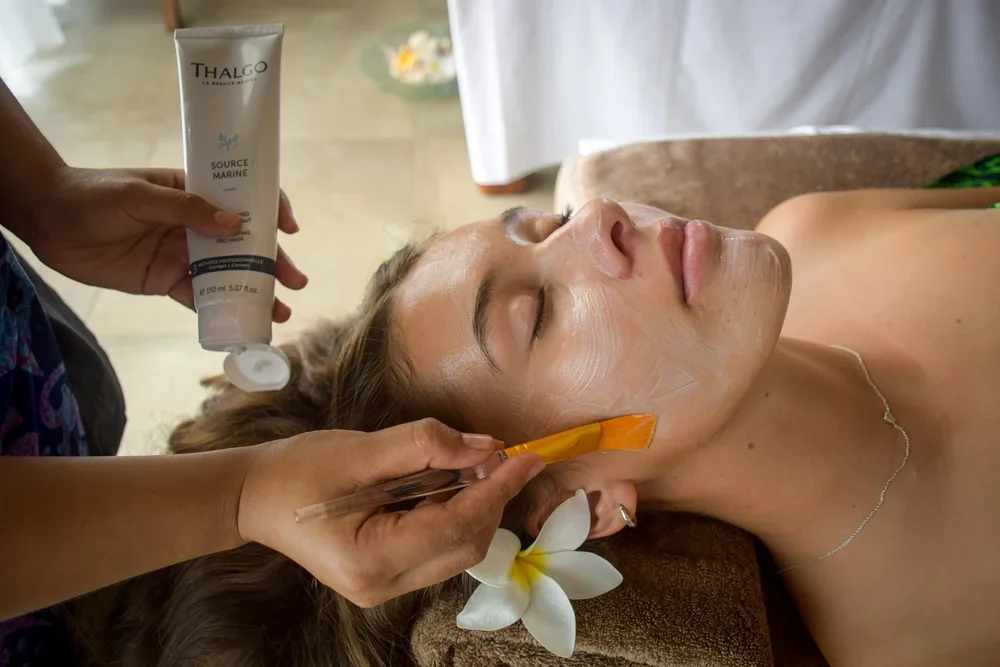 A woman receives a facial treatment with a brush and cream at a spa, with a flower near her ear.