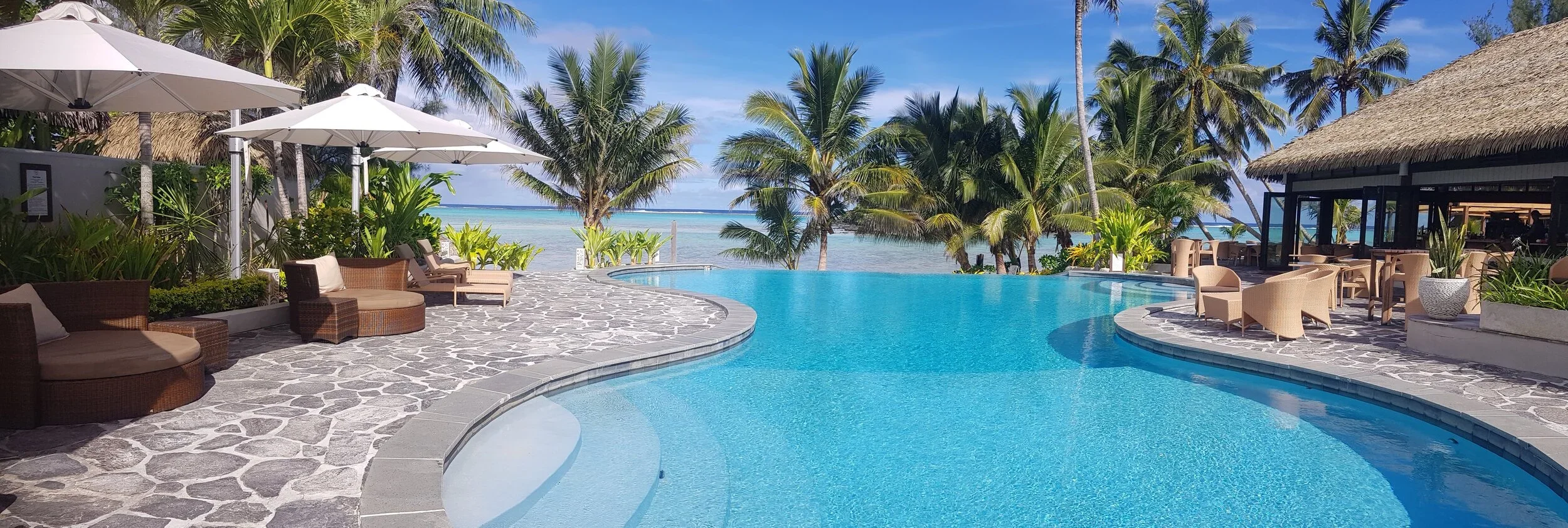A luxurious tropical pool area overlooking the ocean, with lounge chairs, umbrellas, and lush palm trees.