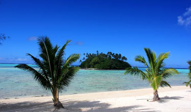 Tropical beach with white sand, palm trees, clear blue water, and a small island in the distance under a bright blue sky.