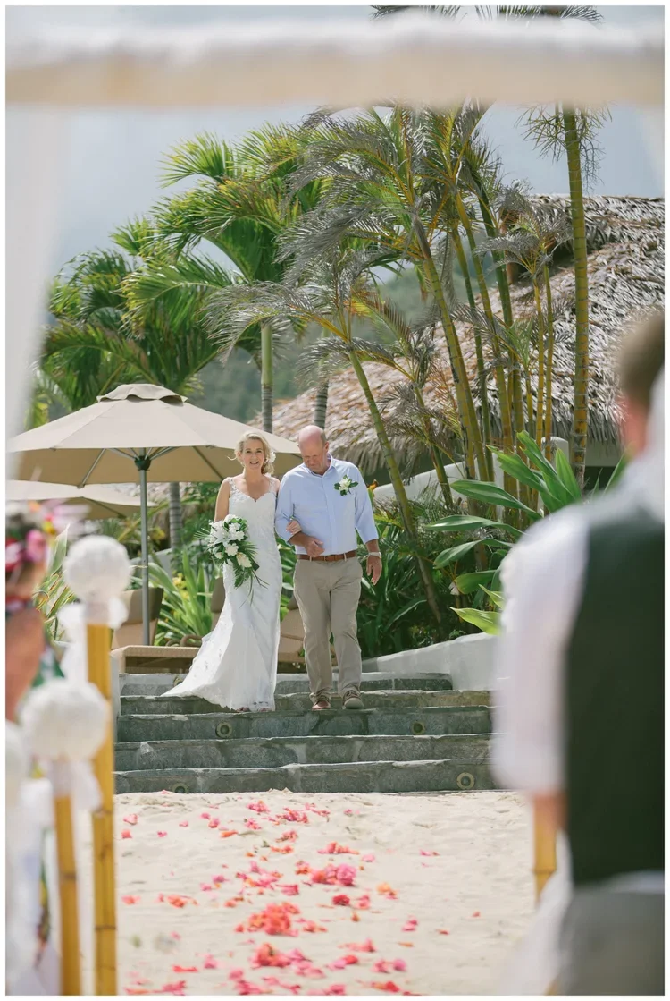 Bride walking down the aisle with her father during a beach wedding ceremony, surrounded by tropical plants and flowers.