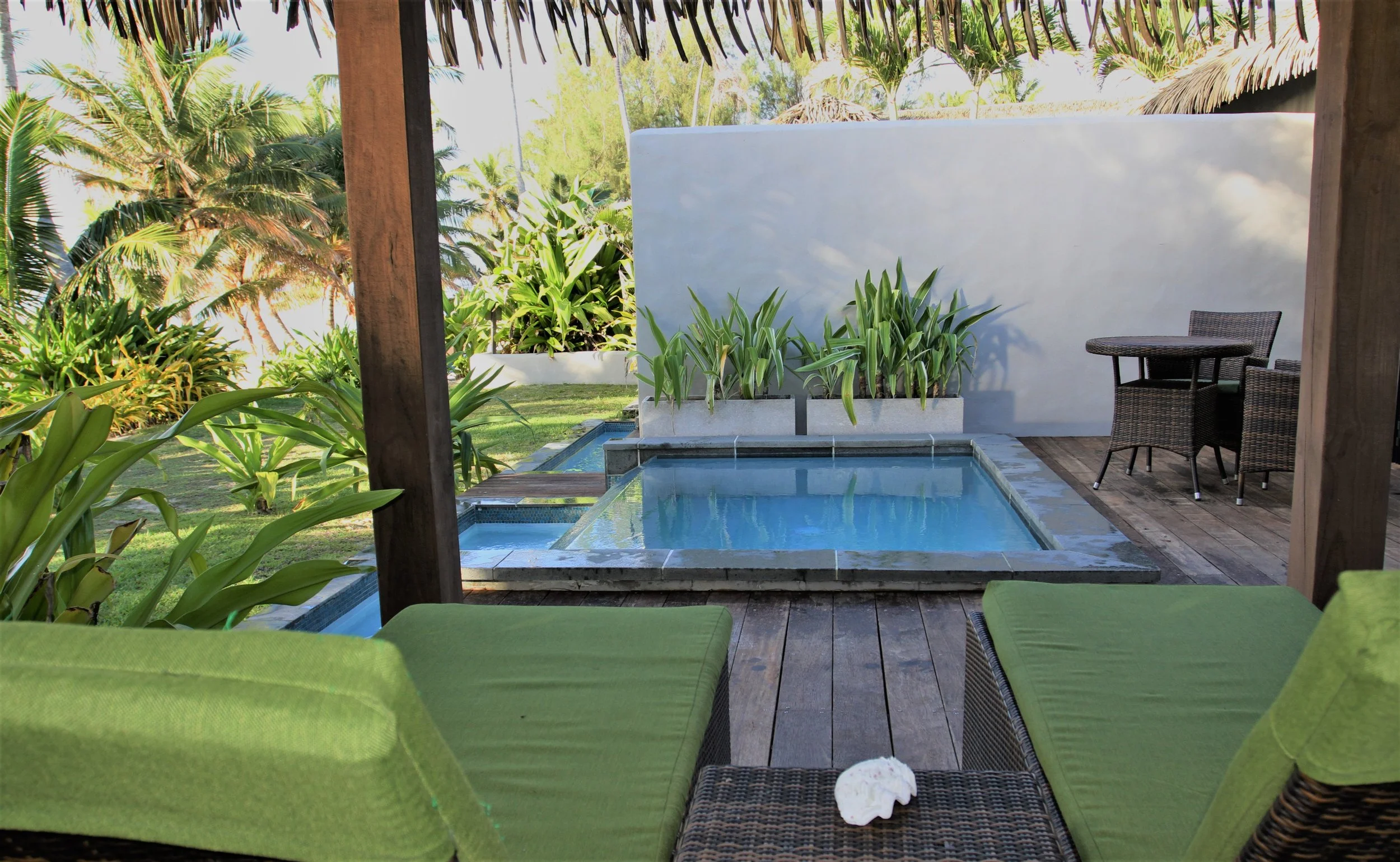 A view from a outdoor patio with two green cushioned lounge chairs, a small woven table, a round table with chairs, a hot tub, lush tropical plants, and palm trees in the background.