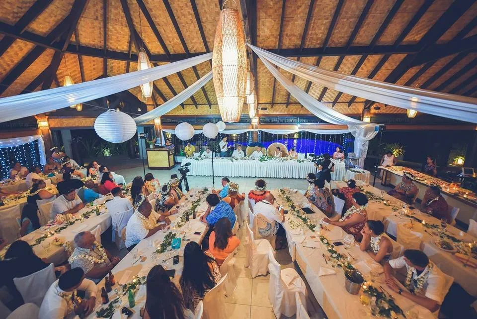 A large wedding reception hall decorated with white drapes and lanterns, with many guests seated at long tables adorned with floral centerpieces, and a head table with the wedding party.