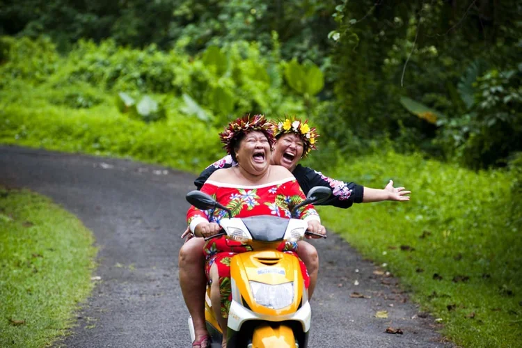 Two women wearing colorful floral outfits and flower crowns riding a yellow scooter on a forested path, laughing and enjoying the moment.