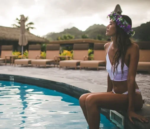 A woman sitting by a swimming pool outdoors, wearing a floral crown and swimsuit, with lounge chairs and umbrellas in the background.