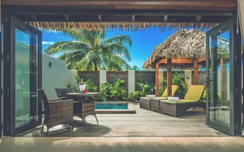 View of a tropical outdoor patio with lounge chairs, a dining table, palm trees, and a thatched-roof tiki hut under a clear blue sky
