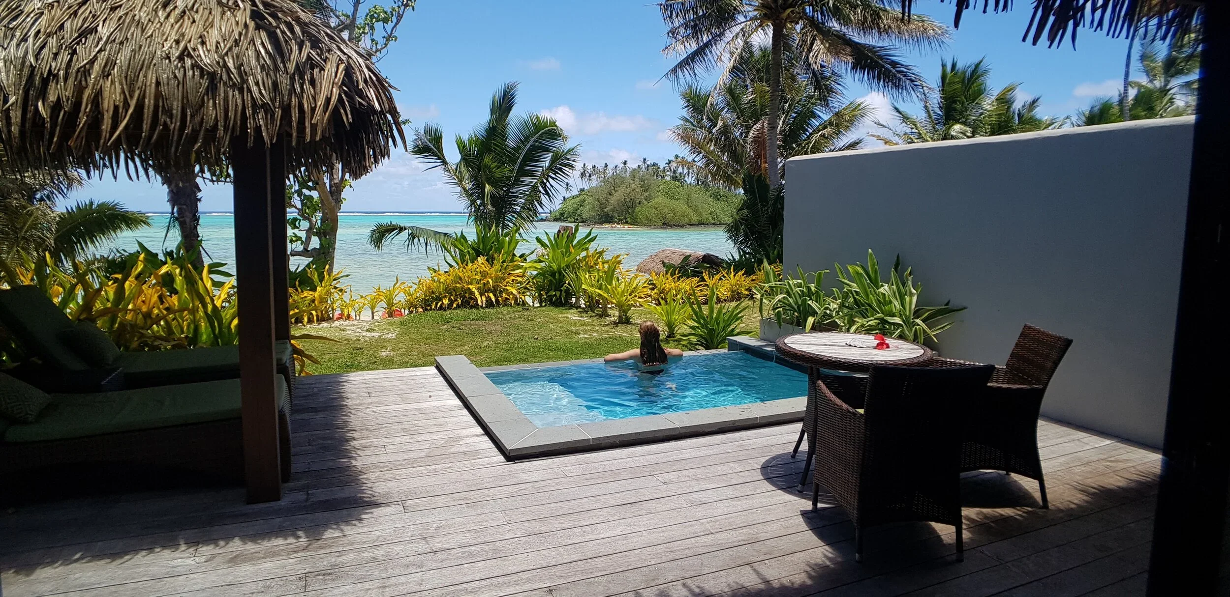 A person relaxing in a small pool on a patio overlooking a tropical beach with palm trees and blue ocean
