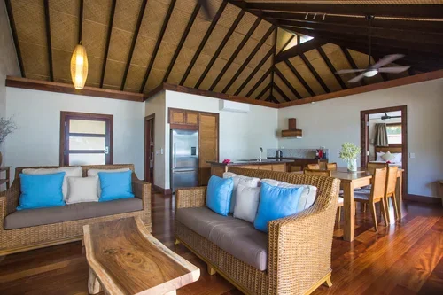 Living room with wicker sofas, blue and white pillows, wooden coffee table, kitchen with refrigerator and sink, and dining area with wooden table and chairs in a tropical-style open layout.