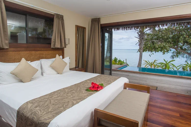 A bedroom with a large bed, beige pillows, and a beige runner, overlooking an ocean view through large sliding glass doors, with a small pool or hot tub outside and tropical trees.