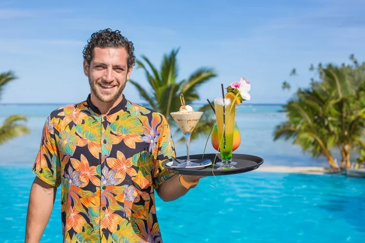 A man smiling at a tropical poolside, holding a tray with three colorful cocktails, with palm trees and the ocean in the background.