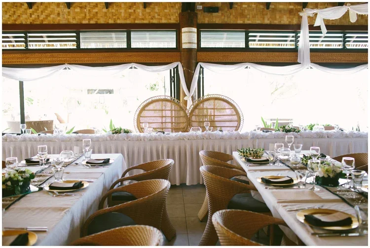 Decorated banquet hall with round tables set for a formal event, white tablecloths, gold and black napkins, wine glasses, floral centerpieces, and a window with drapes in the background.
