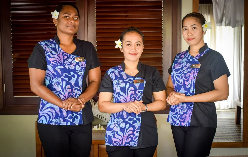 Three women in matching black and blue floral uniforms with flower hair accessories standing indoors, smiling, in front of wooden window shutters.