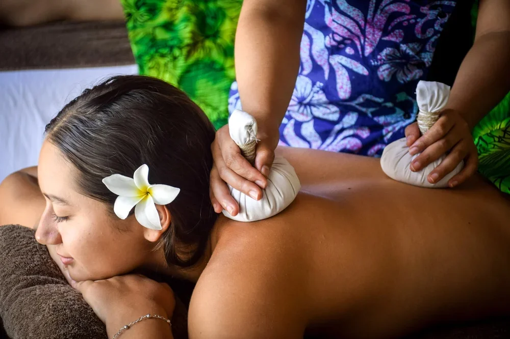 A woman receives a massage with herbal compresses at a spa, with a flower in her hair and colorful attire of the masseuse.