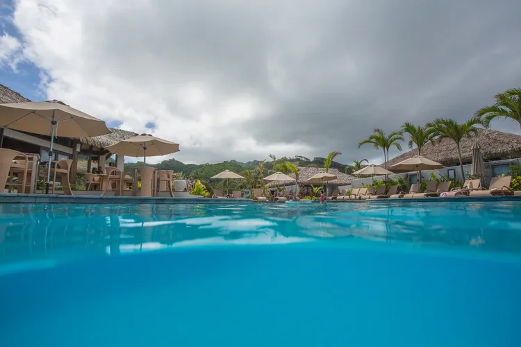 View of a resort swimming pool with umbrellas, lounge chairs, palm trees, and a cloudy sky.