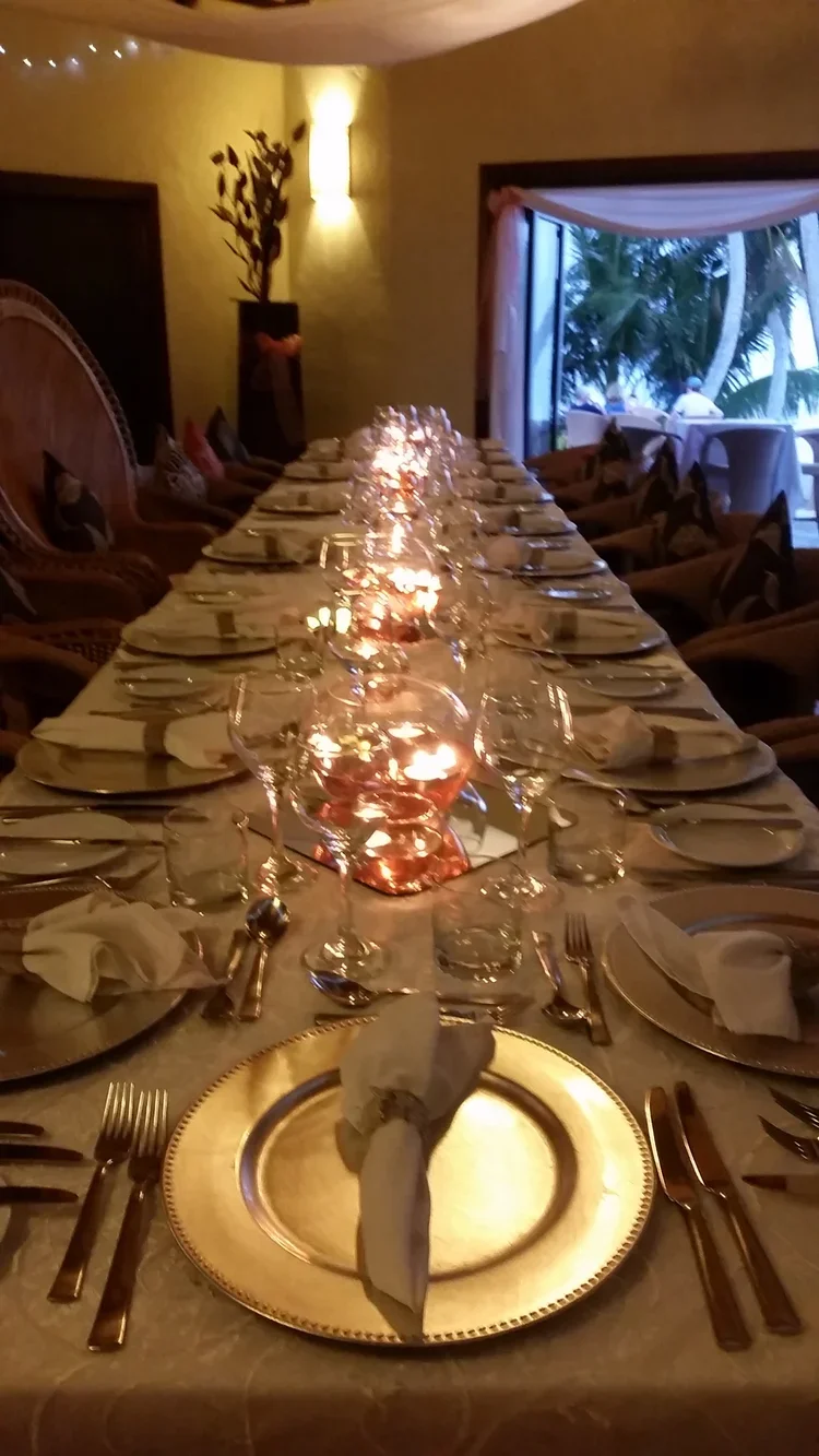 A long dining table set with plates, silverware, and glasses, adorned with candles inside glass holders, in a warmly lit room with a window showing palm trees outside.