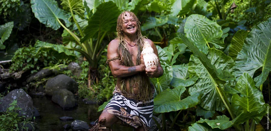 A smiling man with long dreadlocks, wearing a striped skirt, holding a conch shell while standing among large green tropical leaves.