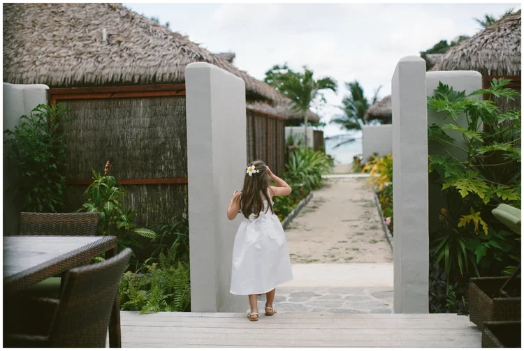 A young girl in a white dress with a flower in her hair walks through an outdoor passageway toward a beach, surrounded by tropical plants and thatched-roof buildings.