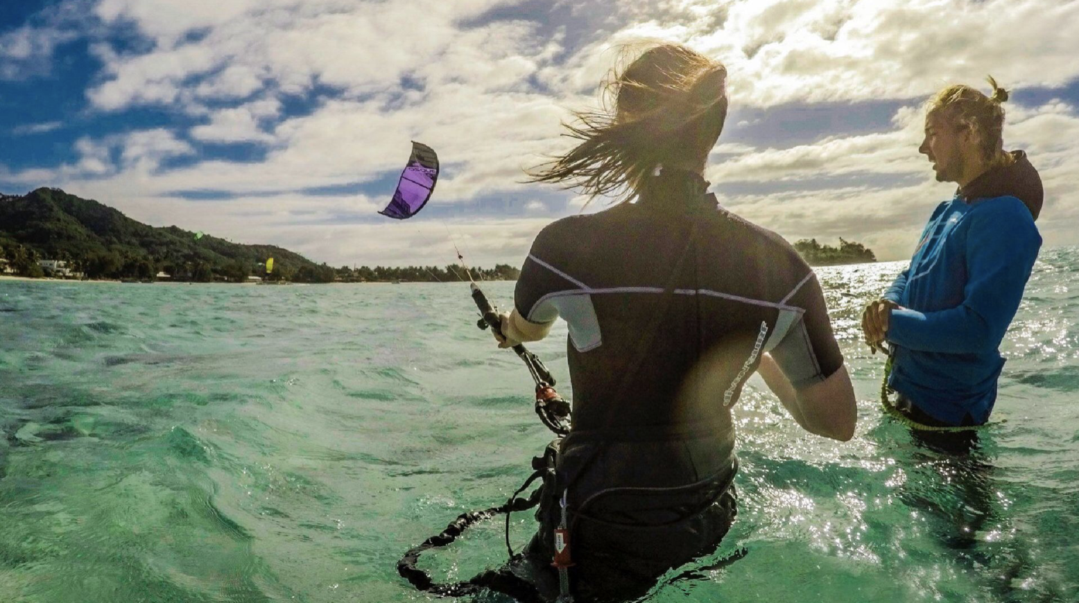 Two people stand in the water, with one holding a kite surfing control bar, while the other talks to them. The sun is shining brightly, and a kite is flying in the sky over a coastal landscape with hills and trees.