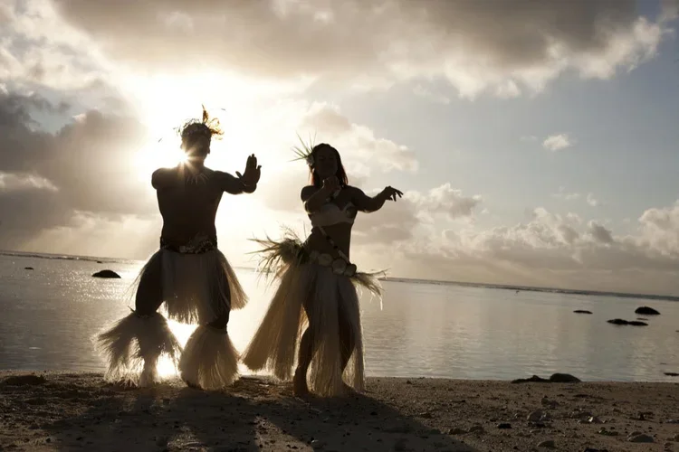 Two people wearing traditional Polynesian hula costumes dancing on a beach at sunset.
