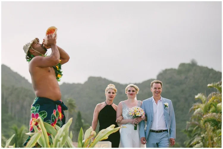 A shirtless man wearing a flower necklace and colorful shorts is playing a conch shell at an outdoor wedding ceremony, with three people standing behind him against a backdrop of green hills.