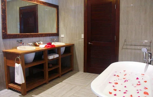 A bathroom with double sinks and a large mirror above, a wooden vanity, and a bathtub filled with water and rose petals.
