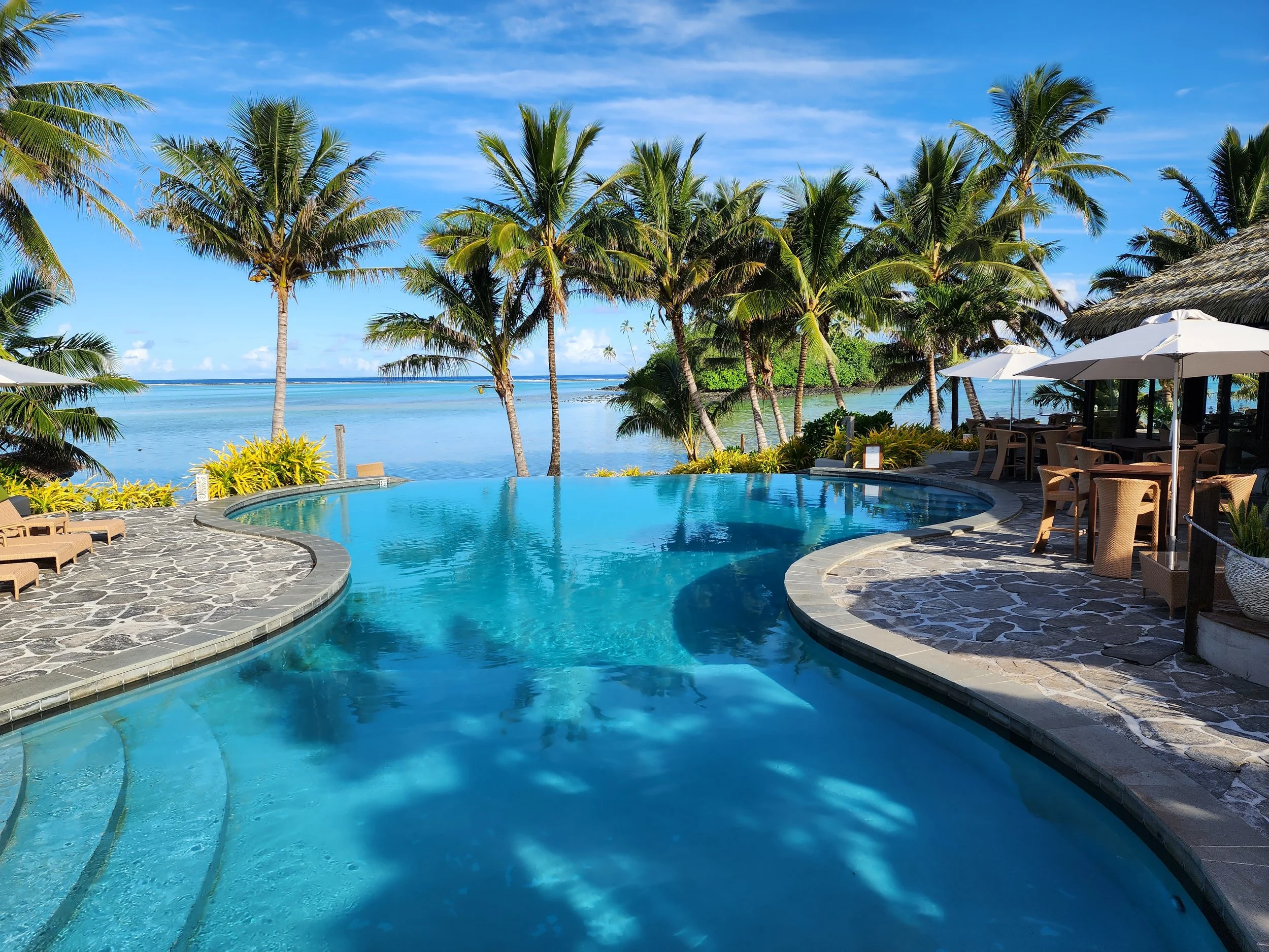A tropical poolside scene with a curved swimming pool, palm trees, and a view of the ocean in the background. There are lounge chairs and tables with umbrellas on a stone patio.