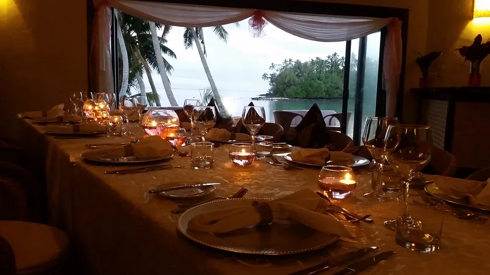 A dining table set for a meal with napkins, glassware, plates, and candles, overlooking a tropical beach with palm trees through a large sliding glass door.