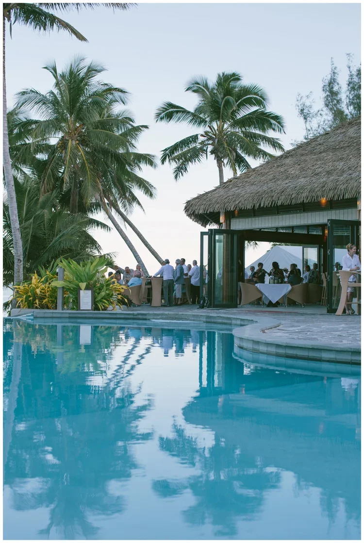 A tropical outdoor restaurant overlooking a swimming pool with palm trees and guests dining.