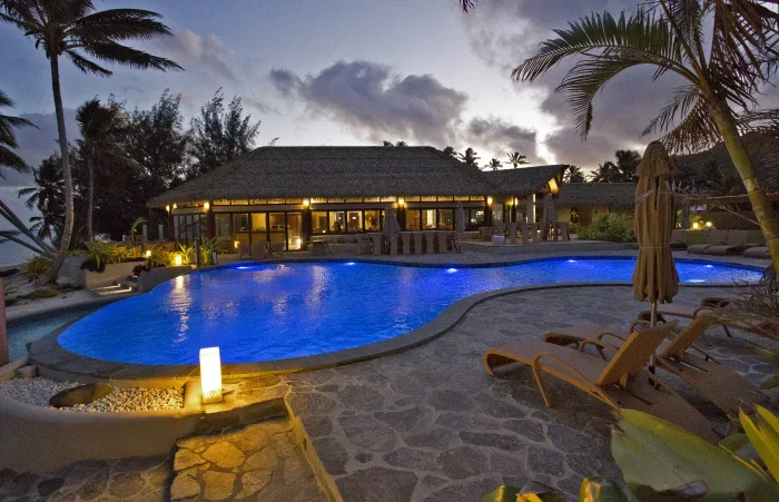 A tropical resort pool area at sunset with lounge chairs, palm trees, and a thatched-roof building in the background.