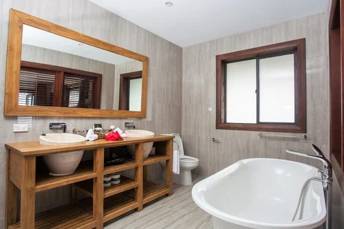 Bathroom with wooden vanity, large mirror, sink, toilet, and bathtub, window with wooden trim, neutral-colored walls and flooring.