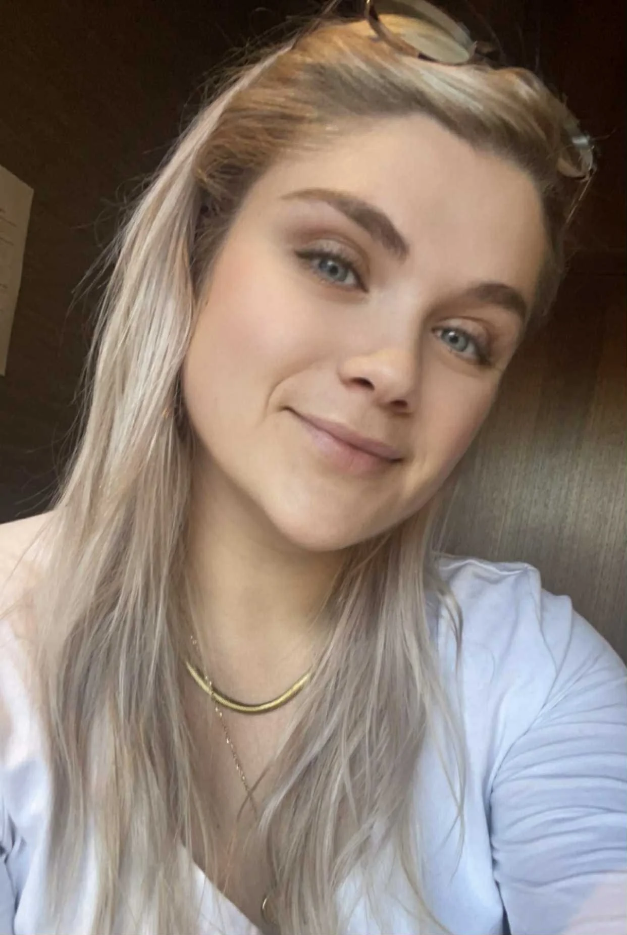 A young woman with long blonde hair, wearing glasses on her head, a white shirt, and layered gold necklaces, smiling at the camera.