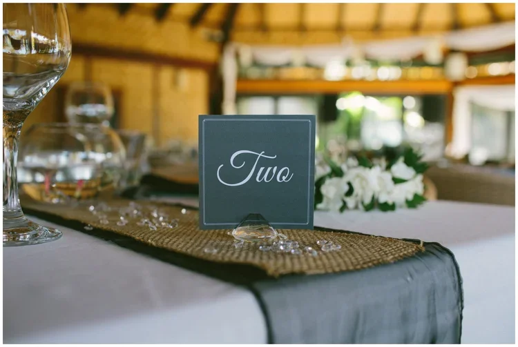 Table setting with a black card labeled 'Two', a wine glass, and a floral centerpiece in a rustic venue.