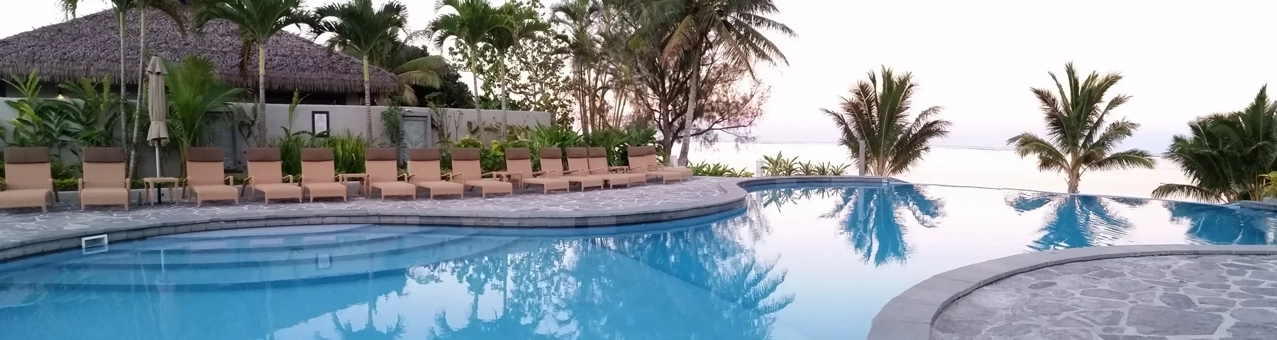 Poolside view at sunset with lounge chairs, umbrellas, palm trees, and a thatched-roof hut in a tropical setting.