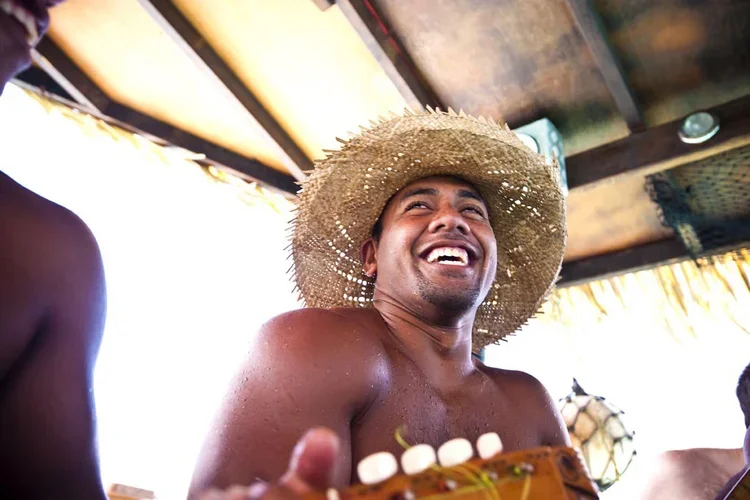 Man with a wide-brimmed straw hat smiling and laughing at a beach or tropical setting.