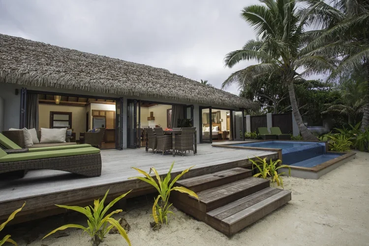 Beachside villa with wooden deck, outdoor dining area, lounge chairs, a pool, and palm trees.