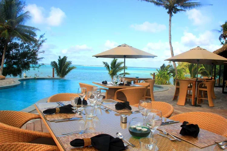 Outdoor restaurant setup overlooking a pool and ocean with wicker chairs, tables with glassware, napkins, and umbrellas, surrounded by palm trees under a blue sky.