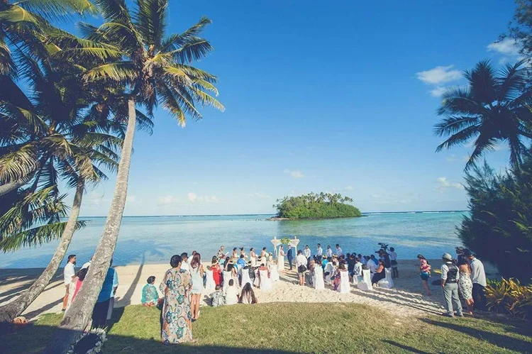 A beach wedding ceremony with guests seated on white chairs facing the water, with palm trees on either side and a small island in the distance under a blue sky.