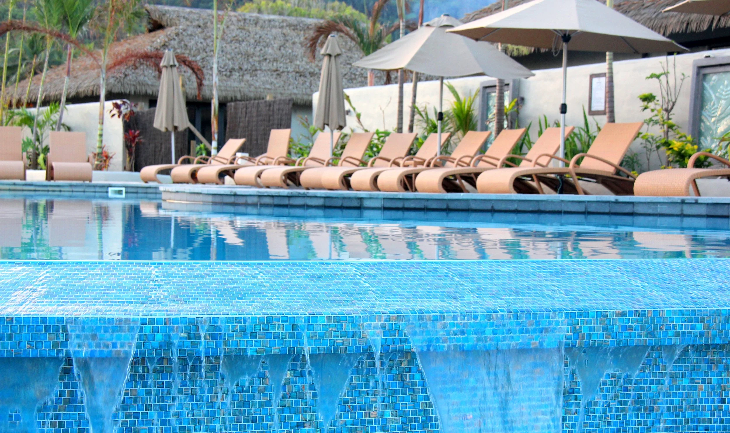 A swimming pool with a waterfall feature in the foreground, lined with multiple beige lounge chairs and umbrellas, surrounded by tropical plants and thatched-roof structures.