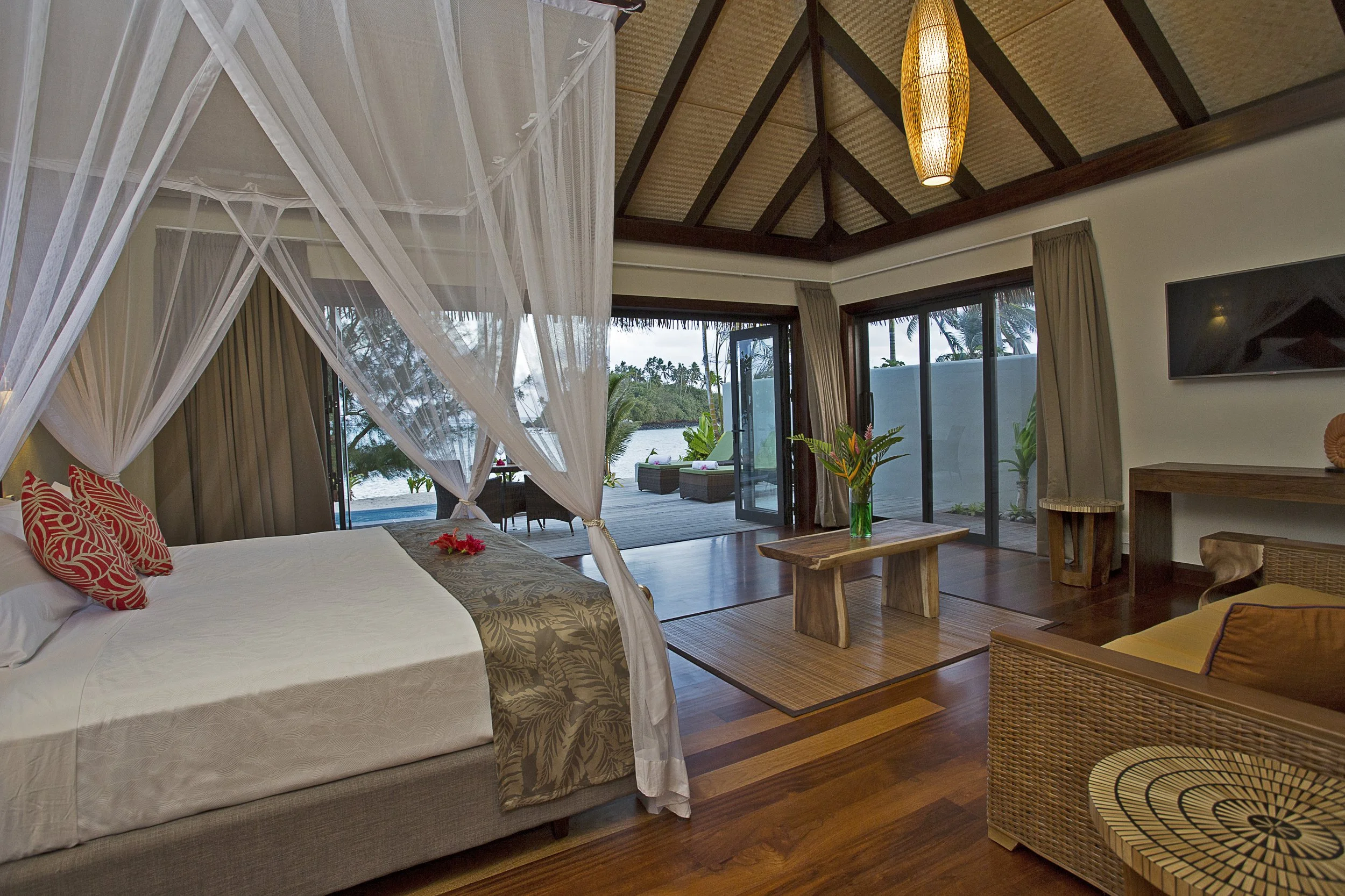 A luxurious bedroom with a mosquito net canopy over the bed, armchair, wooden coffee table with a plant, sliding glass doors opening to a tropical outdoor space with a view of water and palm trees.