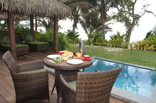Outdoor poolside dining setup with a round table, four wicker chairs, a tray of food, cups, and drinks, surrounded by palm trees and lush greenery.