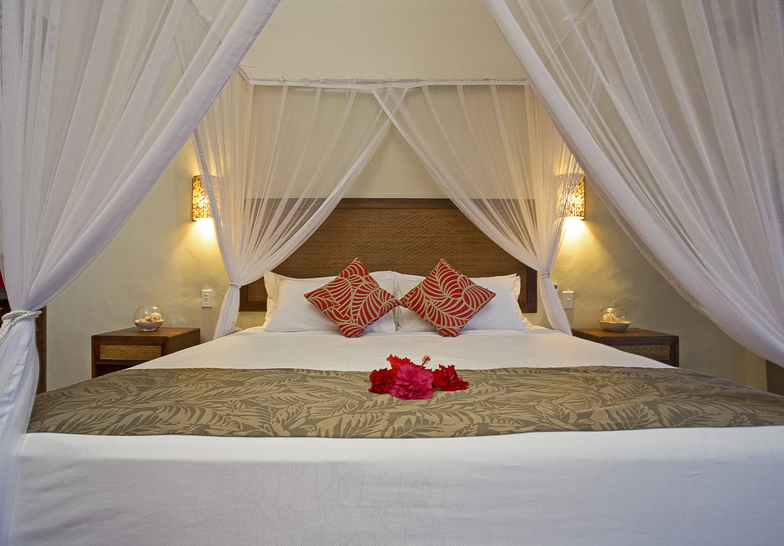 A hotel bed with white sheets, decorative red and white patterned pillows, and a floral arrangement on the bed. The bed is enclosed with white mosquito netting and has wooden nightstands with lamps on each side.