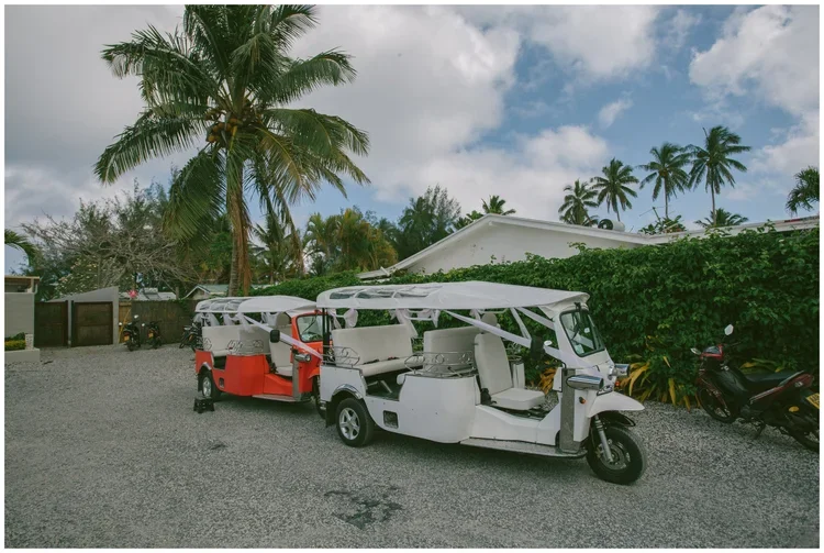 Two tuk-tuks parked on gravel in front of palm trees and a white house with green hedge, cloudy sky overhead.