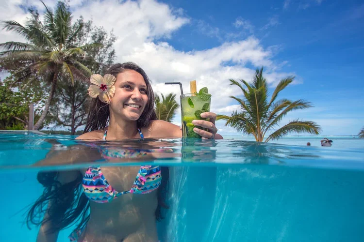 Woman smiling with a flower in her hair holding a mint lime cocktail in a pool with palm trees and a blue sky in the background.