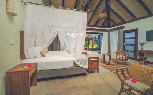 A tropical bedroom with a four-poster bed with white mosquito netting, wooden furniture, a sitting area, and open doors to a patio with greenery.