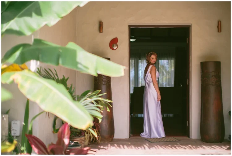 A woman in a long, elegant white dress standing in a doorway, facing towards the camera, in a room with tall, brown vases, indoor plants, and natural light.