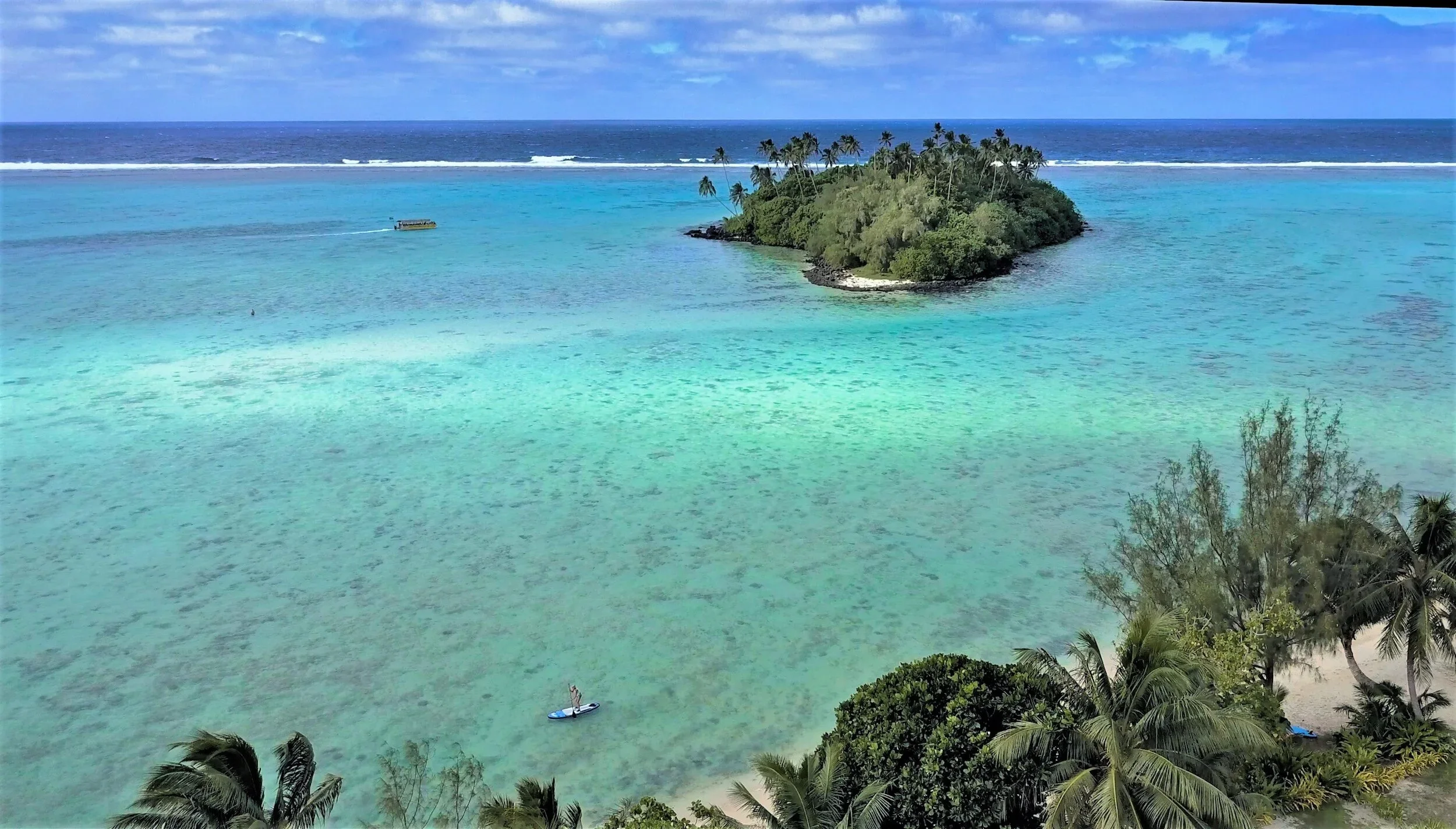 Tropical island surrounded by turquoise waters with a small boat and a paddleboarder, lush palm trees, and a partly cloudy sky.