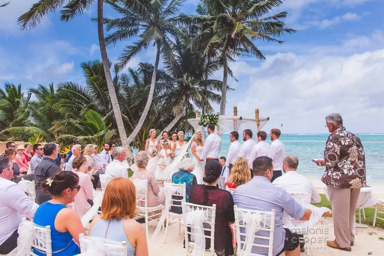 A beach wedding ceremony with guests seated on white chairs, palm trees, and the ocean in the background.