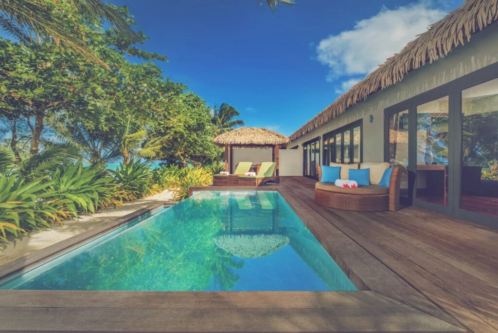 A luxurious outdoor pool area at a tropical resort with lounge chairs, a thatched gazebo, and a modern building with large glass doors, surrounded by lush green plants and trees under a partly cloudy blue sky