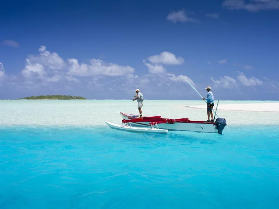 Two men fishing from a small boat near a sandy beach with clear blue water and a partly cloudy sky.