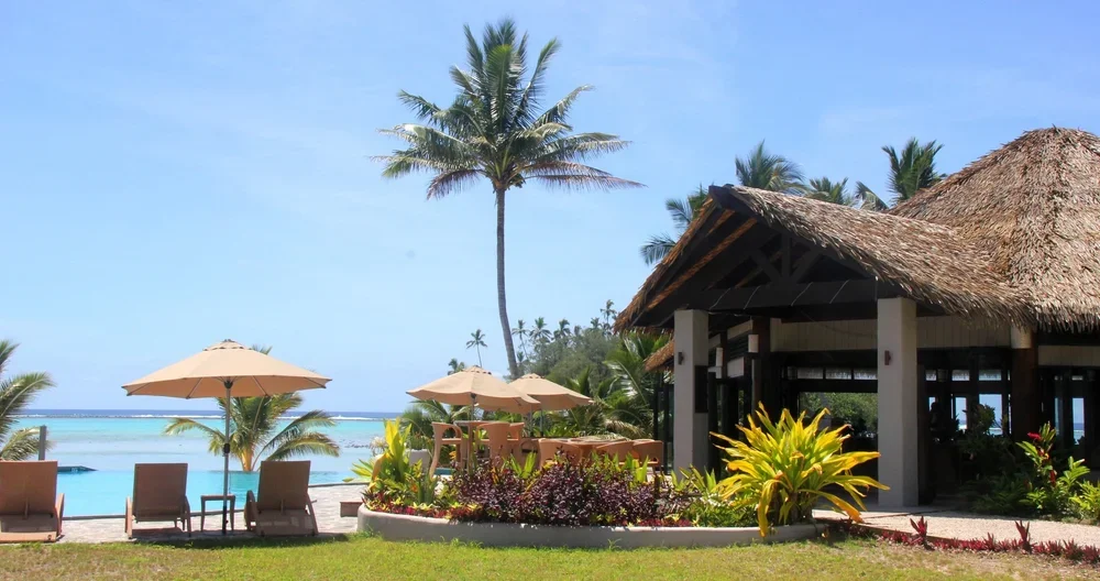 A tropical beachside resort with palm trees, a swimming pool, thatched roof hut, lounge chairs, and umbrellas under a blue sky.