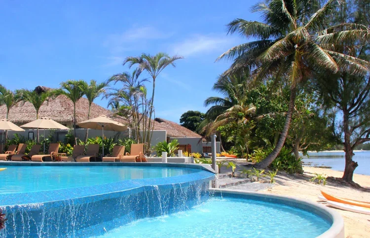 Swimming pool with waterfall feature, surrounded by palm trees, lounge chairs, umbrellas, and a beach in the background on a sunny day.